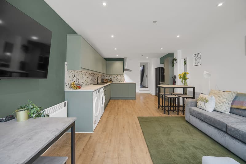 Open-plan kitchen and living room in a Cardiff HMO with sage green units, oak floor and grey sofa