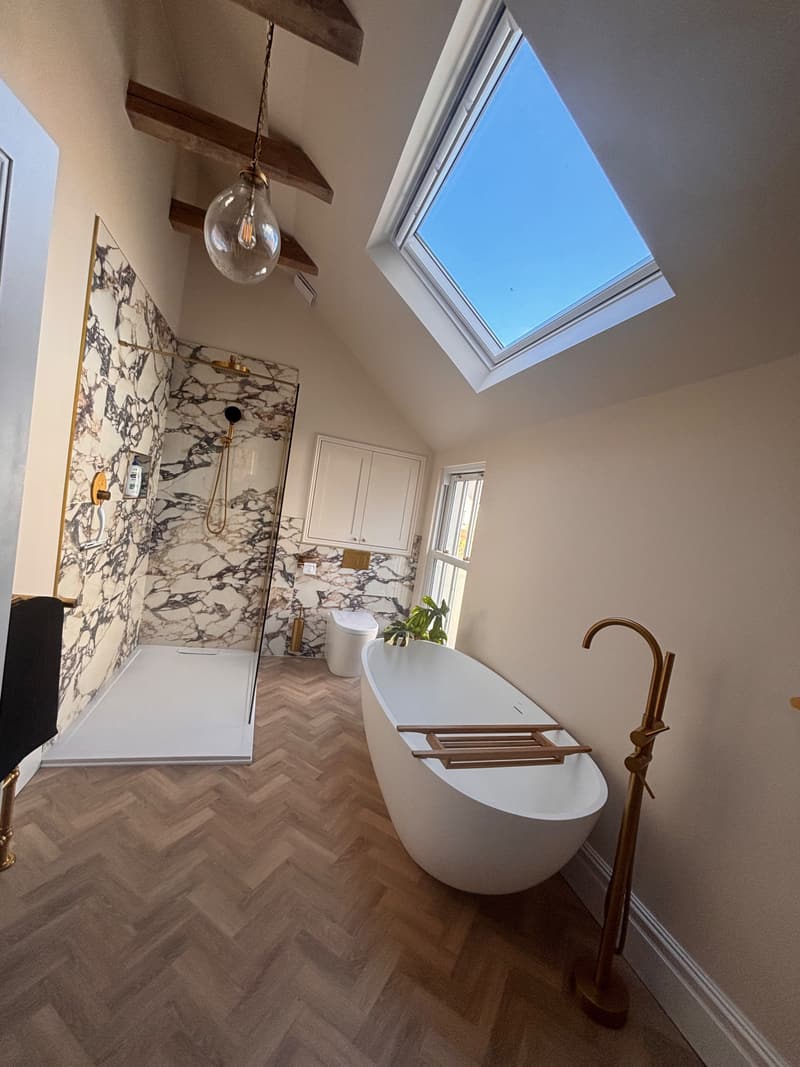 Vaulted loft bathroom with chunky timber beams, marble-effect walls, floor-standing brass bath filler and herringbone floor